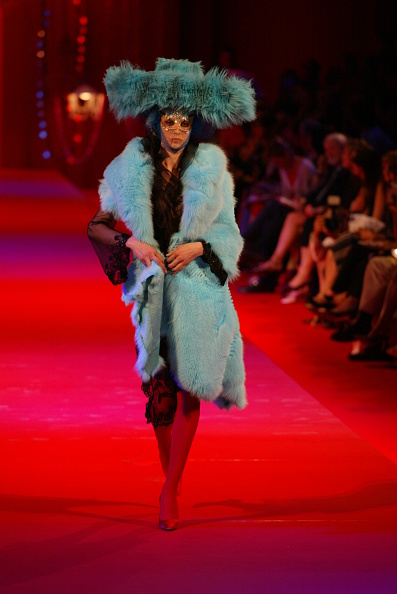 A fashion model is pictured walking down the runway for the Christian Lacroix Fall 2002 show, showcasing a fluffy faux fur coat and a wide brim