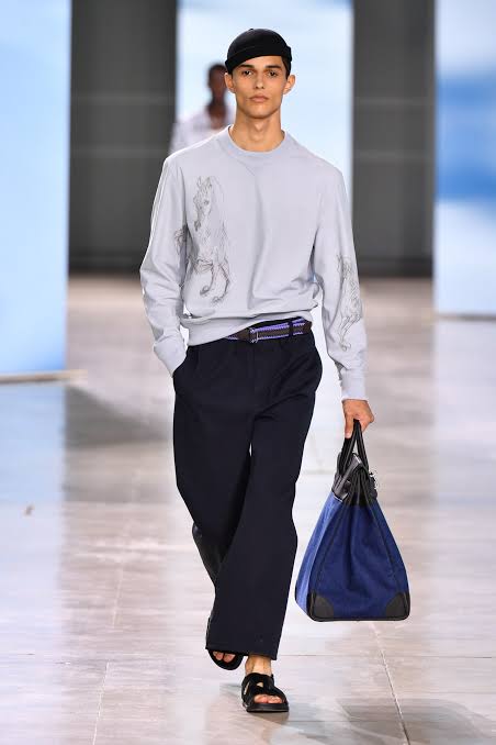 Model wearing gray long sleeve tee shirt, black pants and blue bag at the Paris Fashion Week Men’s spring 2025
