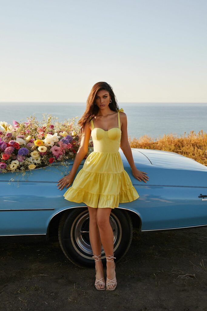 dark haired model wearing Butter Yellow dress leaning on car