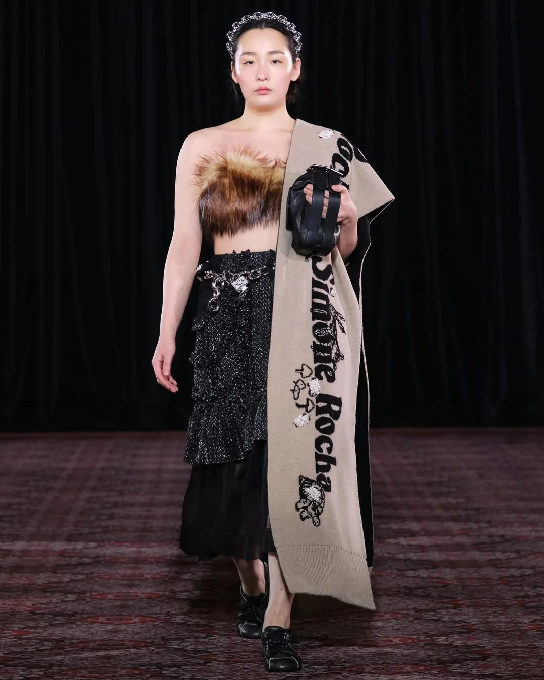 Min-ha Kim is shown walking down the catwalk at the Simone Rocha Fall 2025 fashion show, dressed in a fur-trimmed top, a layered skirt with a mesh design, and a large scarf thrown over one shoulder, similar to Jenna Ortega’s look on the “Wednesday” Season 2 Seoul press tour.