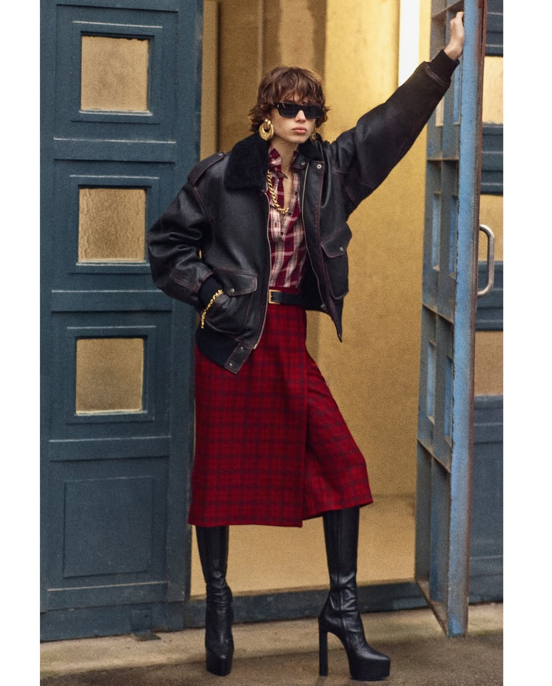 Mathilda Gvarliani models the Saint Laurent Pre-Fall 2025 collection in a black bomber jacket with a shearling collar, a plaid shirt, and a wrap midi skirt