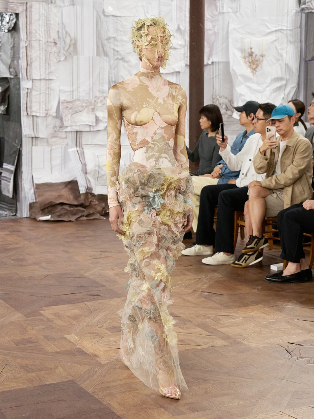 Pien Laureijnssen walks down the runway for Maison Margiela’s Artisanal 2025 presentation, dressed in a long-sleeve turtleneck gown, a skin-tight bodysuit with a morte flower motif, and a tulle illusion maxi skirt with lace appliqué decorations.