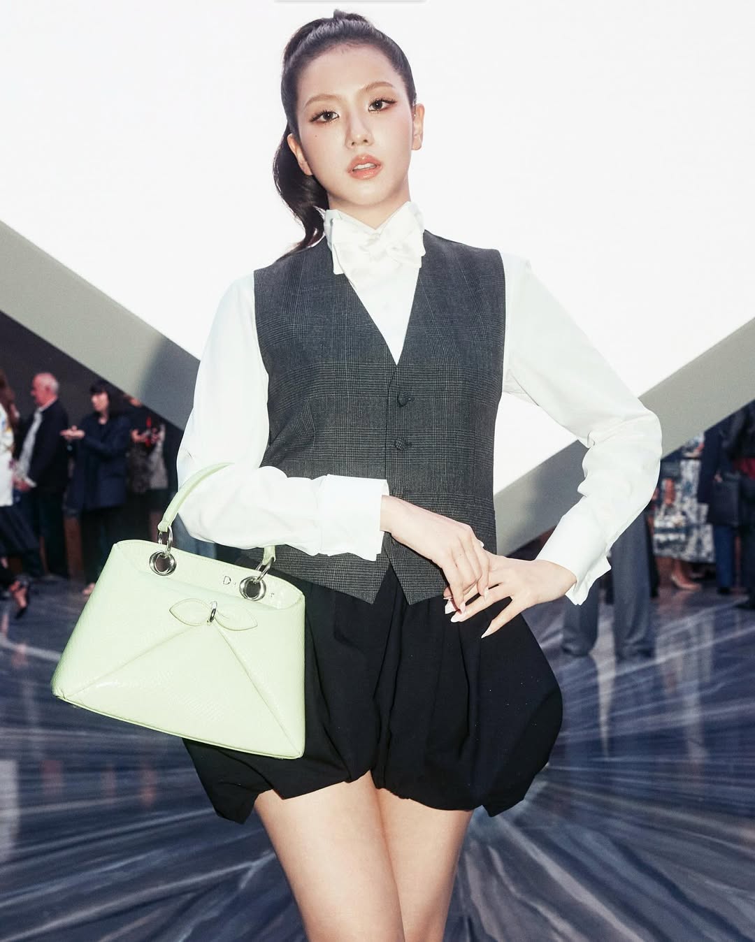 BLACKPINK Jisoo poses for photos during the Christian Dior Women’s Wear Spring/Summer 2026 show, wearing a gray vest, a white blouse with a matching bow tie, a black pleated bubble mini skirt, black slingback sandals, and a mint green handbag.