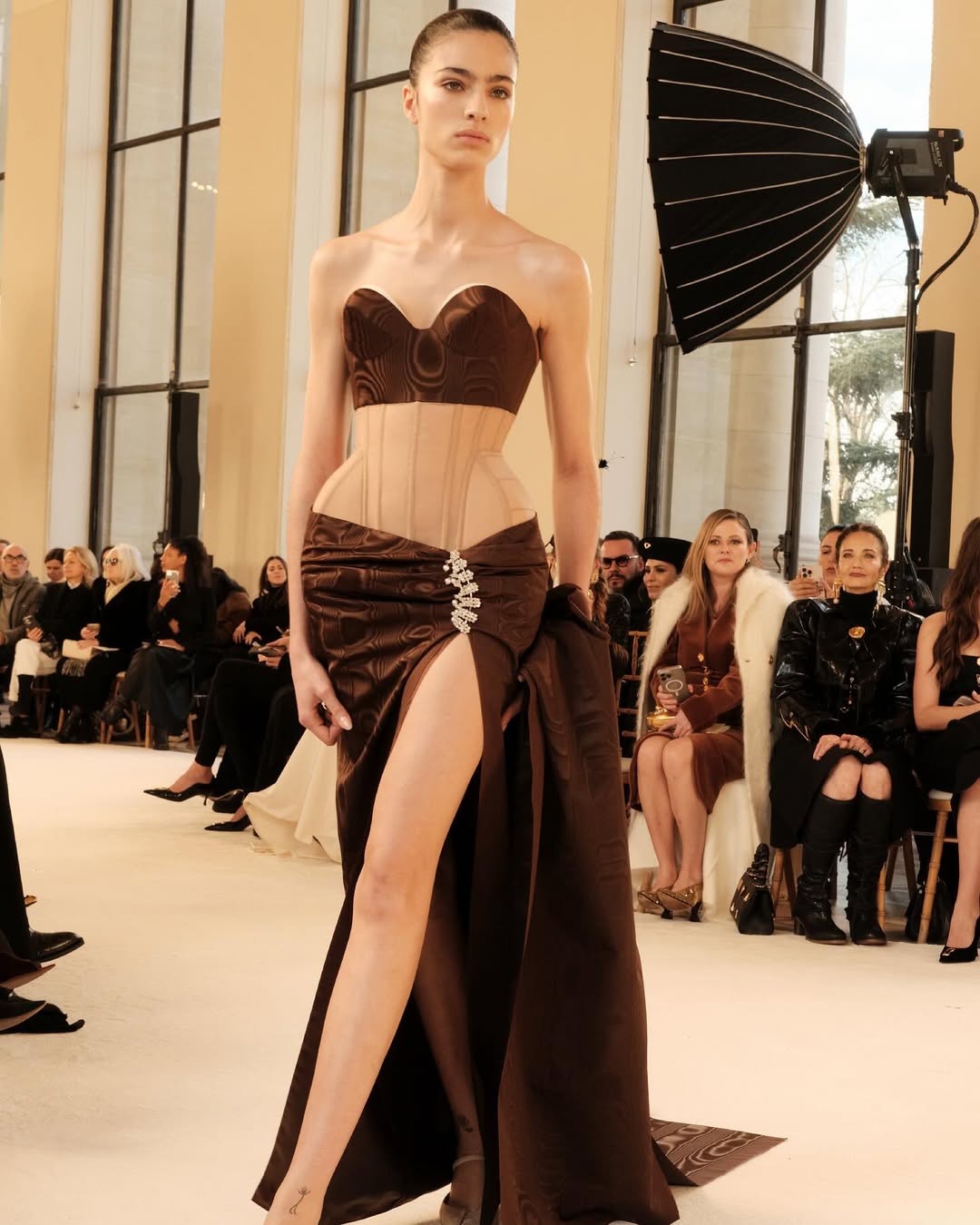 Loli Bahia walks the Schiaparelli Haute Couture Spring/Summer 2025 runway in a bustier dress with a sweetheart neckline of brown moiré vintage ribbons and a sheer mesh corset with exaggerated hips and a slit secured with an antique rhinestone pin.