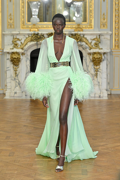 Ajak Dhieu walks down the runway for the Tamara Ralph Haute Couture Spring/Summer 2024 show, dressed in a green tea-draped chiffon gown with voluminous sleeves decorated with ostrich feathers and a gold metal waist belt.